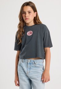 Gray cropped t-shirt in soft cotton, featuring a round neckline and short sleeves, with a pink circular patch on the chest. Light blue jeans with a faded finish.