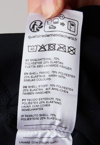 Étiquette d'entretien en tissu noir affichant les instructions de lavage et la composition du matériau : 75 % polyester, 25 % élasthanne ; texte et symboles multilingues.