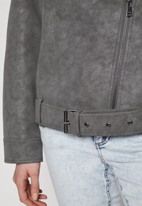 Veste en daim gris avec une fermeture éclair à l'avant, une taille ceinturée avec une boucle en métal et une texture douce ; associée à un jean en denim clair.