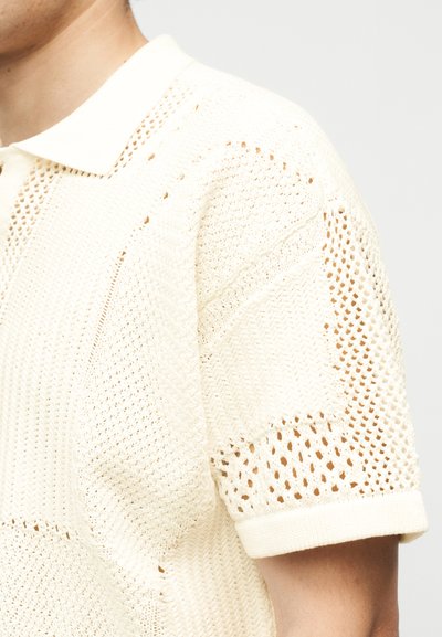 Κοντινό πλάνο ενός ατόμου που φοράει ένα πλεκτό πόλο σε κρεμ απόχρωση με διάφορα ανοιχτά σχέδια και κοντά μανίκια.