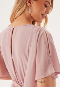 Lichtroze, vloeiende jurk met korte mouwen en een uitsparing op de rug. Gladde, zachte stof met een geknoopte taille, geaccentueerd door eenvoudige oorbellen.