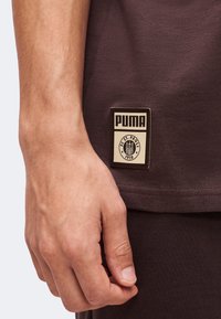 Κοντινό ενός χεριού που ακουμπά δίπλα σε σκούρο καφέ πουκάμισο με μπεζ και μαύρο ταμπελάκι λογοτύπου PUMA και FC St. Pauli 1910.