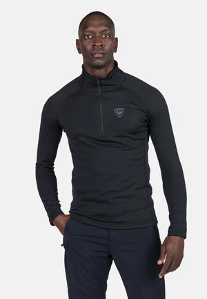 Sudadera negra de manga larga, con cremallera de cuarto y un patrón texturizado, que presenta un pequeño logo en el pecho. La tela parece ajustada y suave.