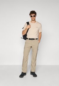 Beige bomull T-skjorte med logo, beige cargo-bukser, svart belte, svarte tursko, og lyse solbriller. Ryggsekk slengt over skulderen.