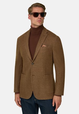 Homme portant un blazer marron en chevrons sur un col roulé marron foncé, jean bleu et lunettes de soleil foncées, debout devant un fond clair uni.