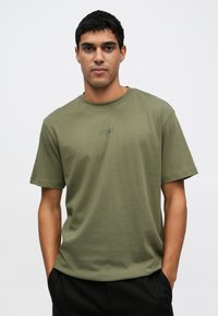 Roheline puuvillane ümara kaelusega T-särk lühikeste varrukatega. Oskab tikitud "BOSS" logo samas toonis. Modell seisab käed taskutes.