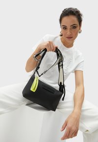 Czarna torebka crossbody, wykonana z gładkiego materiału, z kontrastową, neonowo-żółtą metką oraz czarnym paskiem w białym wzorze. Prostokątny kształt.