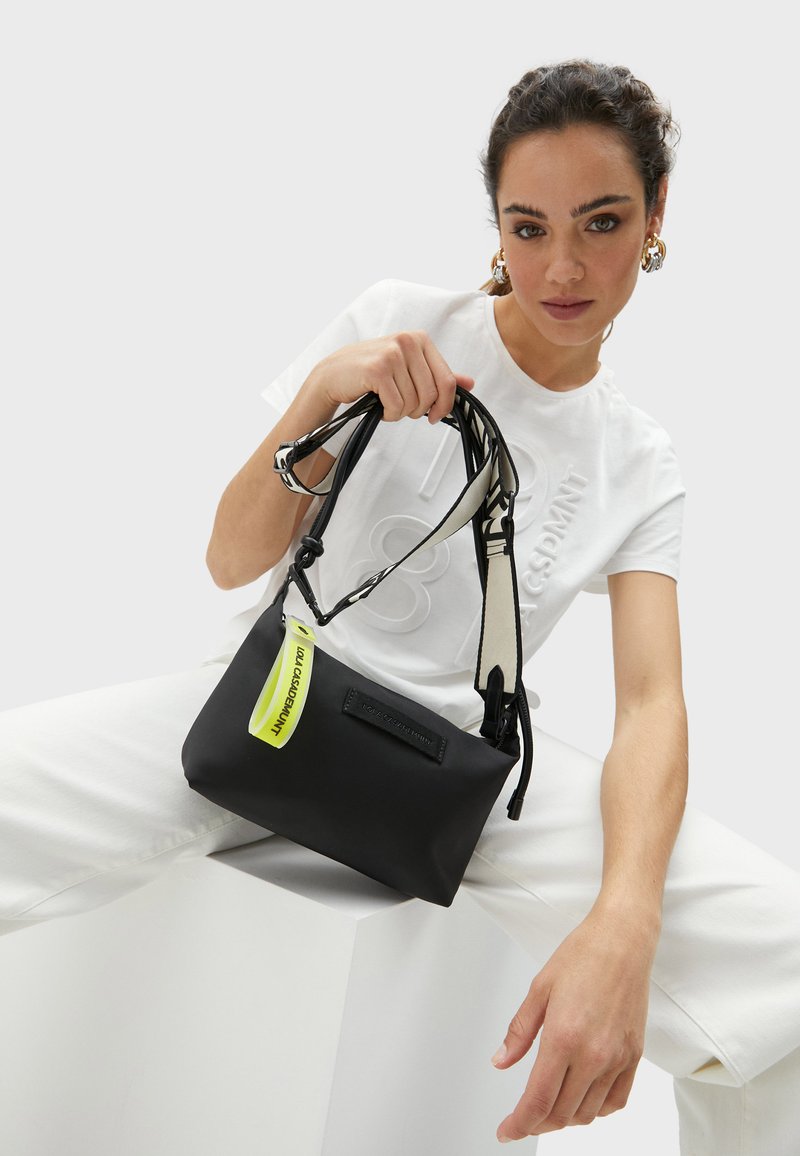 Czarna torebka crossbody, wykonana z gładkiego materiału, z kontrastową, neonowo-żółtą metką oraz czarnym paskiem w białym wzorze. Prostokątny kształt.