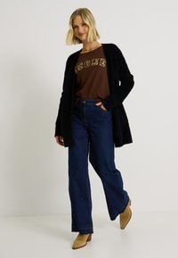 Cardigan noir en peluche, tee-shirt graphique marron avec lettrage léopard, et jean ample bleu foncé associé à des bottines à talons beiges.