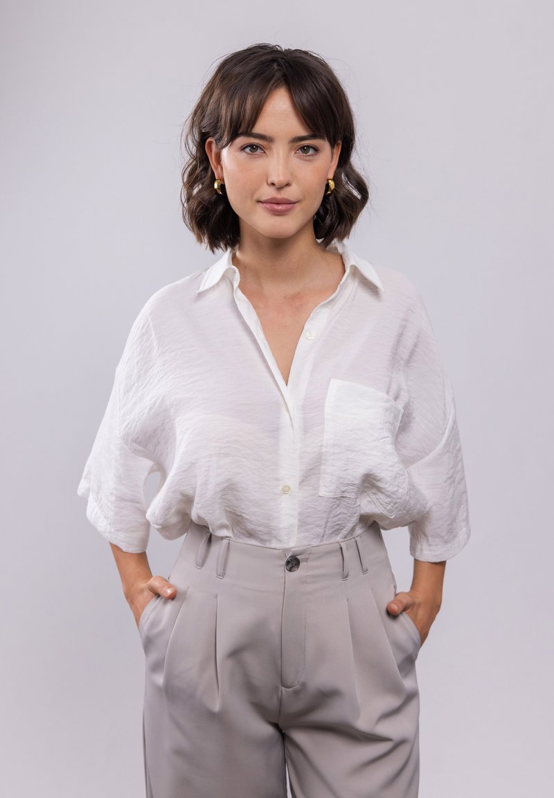 Weißes, leichtes Hemd mit Knopfverschluss und Tasche; kombiniert mit hochtaillierten, gefalteten hellgrauen Hosen. Glatter Stoff, lockere Passform.
