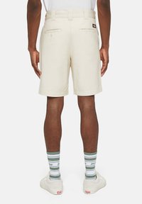 Pantaloni corti beige con vestibilità classica, dotati di tasche posteriori, passanti per cintura e un piccolo logo. Abbinati a calze a righe verdi e bianche.