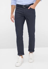 Chinos bleu marine à coupe droite, revers roulés et ceinture tressée texturée, associés à des baskets blanches. Tissu lisse avec des détails subtils.