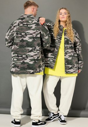 Dos modelos lucen chaquetas de camuflaje a juego, con prendas superiores de color amarillo brillante, pantalones blancos y zapatillas negras; uno está de espaldas y el otro de frente.