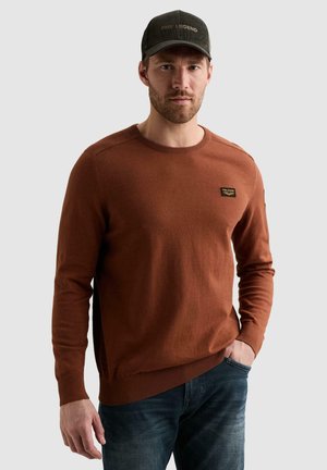 Homme portant un pull marron à manches longues avec un écusson et une casquette foncée, debout avec une main dans sa poche sur un fond uni.