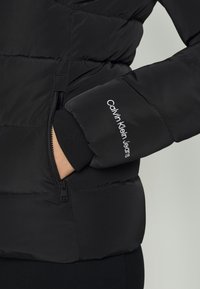 Manche de doudoune noire avec broderie blanche « Calvin Klein Jeans », main dans la poche latérale zippée, poignet intérieur côtelé visible.