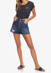 Mørkegrå t-shirt med et guld-logo, parret med højtsiddende denimshorts med knaplukning, frynset kant og slidte detaljer. Sorte sandaler.