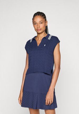 Marineblå strikket ermeløs poloshirt med kabelmønster, hvitstripet krage og sideslister, sammen med et matchende marineblå skjørt med perforeringer.