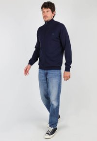 Pull marin zippé avec col montant, en tissu tricoté ; porté avec un jean bleu clair et des baskets noires. Présente un logo discret.