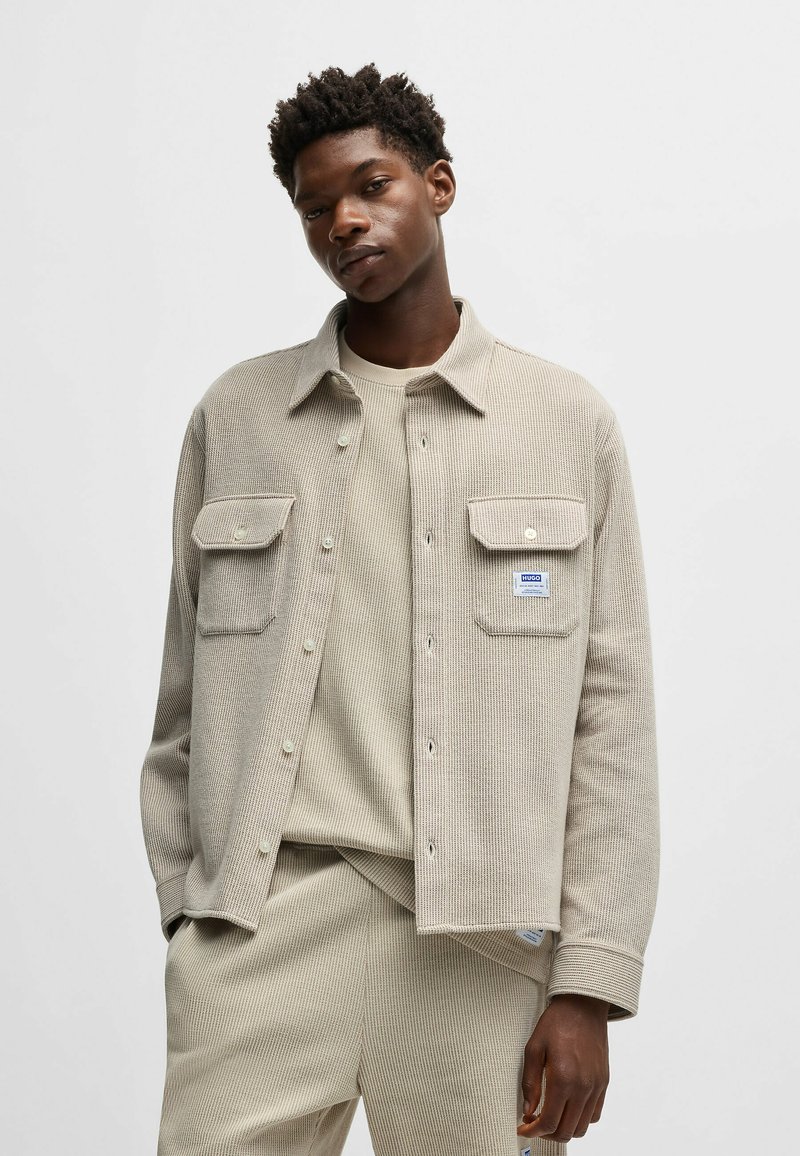 Veste en velours côtelé beige avec deux poches avant, fermeture par bouton et étiquette. Portée sur un pull à col ras du cou beige assorti.