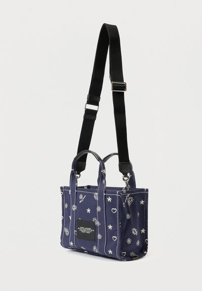 Geantă tote de culoare bleumarin, cu modele albe de flori, stele și inimi, curea de umăr ajustabilă neagră și eticheta neagră cu text.