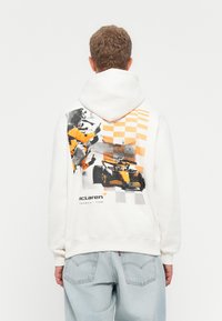 Person iført hvid hoodie med orange og sort grafik, der viser jubelende crowd, racerbiler og ternet flagmønster på ryggen.