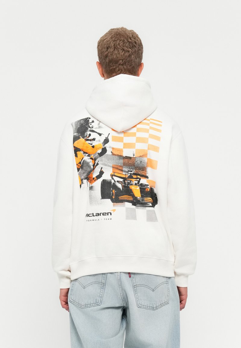 Person iført hvid hoodie med orange og sort grafik, der viser jubelende crowd, racerbiler og ternet flagmønster på ryggen.