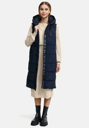 Marineblau gesteppter langer Weste mit Kapuze, ausgestattet mit einem Reißverschluss und Druckknopfverschluss, kombiniert mit einem beigen Kleid und schwarzen Knöchelstiefeln.