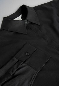 Camisa negra de manga larga con botones, cuello puntiagudo y puños con un solo botón, exhibida plana sobre una superficie clara.
