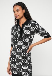 Vestido tipo polo com um padrão preto e branco, mangas curtas e gola preta com fecho de botão. Tecido texturizado com um design gráfico.