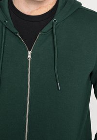 Dark green zip-up hoodie featuring a silver zipper, adjustable drawstrings, and a soft, textured fabric. Includes a black inner layer.