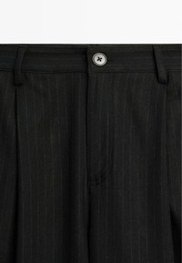 Gros plan sur un pantalon noir à fines rayures, ceinture avec un bouton argenté, passants de ceinture et discrètes rayures verticales.