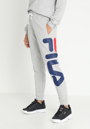 Person iført grå joggingbukser med stort blåt og rødt Fila-logo på benet og sorte Fila-sneakers med hvide såler, stående imod en hvid baggrund.