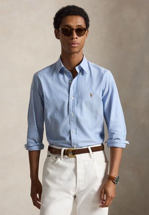 Homme portant des lunettes de soleil marron, une chemise bleu clair boutonnée, un pantalon blanc avec une ceinture marron et une montre-bracelet, debout devant un fond uni.