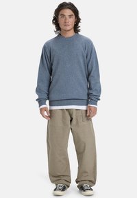 Blauer Strickpullover mit Rundhalsausschnitt, gerippten Bündchen und Saum; kombiniert mit beigen weiten Hosen. Schwarze und weiße Sneakers runden den Look ab.