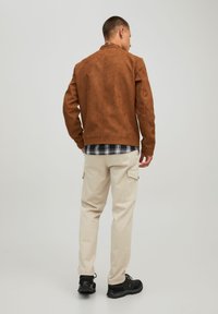 Giacca in suede marrone, pantaloni cargo beige e scarpe nere. La giacca presenta un colletto alto; una camicia a quadri è visibile sotto la giacca.