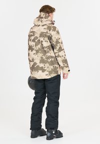 Beige ja ruskeasävyinen kuvioitu lumilautatakki hupulla, yhdistetty mustiin lumihousuihin ja saappaisiin, joissa on mattapinta ja säädettävät osat.