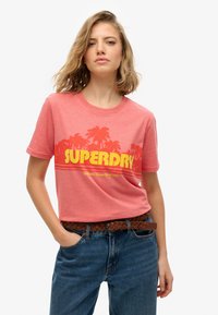 Rød lyng crew-neck t-shirt med gul "SUPERDRY" grafik og palmetema. Matchet med høj talje blå jeans og vævet bælte.