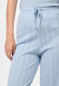 Pogled od blizu na osebo, ki nosi svetlo modre pletene hlače z vertikalnimi črtami in zavezanim pasom, pri čemer leva roka počiva ob njeni strani.