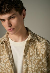 Cămașă bej cu model floral, confecționată dintr-un material texturat, suprapusă peste un tricou crem cu gât rotund, având o margine riglată.