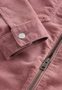 Gros plan sur la manche d'une veste en velours côtelé rose avec un bouton pression argenté et une fermeture éclair visible avec tirette en métal.