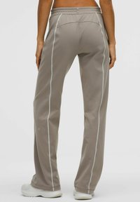 Világosbarna sportnadrág fehér oldalsávokkal, rugalmas derékrésszel és sima, texturált anyaggal; a lábszár alsó részén logó található.