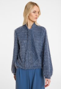 Blauer, flauschiger Pullover mit hohem Kragen und strukturierter Optik. Lange Ärmel und lockere Passform, kombiniert mit weiten blauen Hosen.