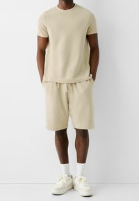 T-shirt en coton beige à manches courtes accompagné d'un short beige. Le modèle porte des baskets blanches et des chaussettes blanches côtelées. Texture lisse, coupe décontractée.