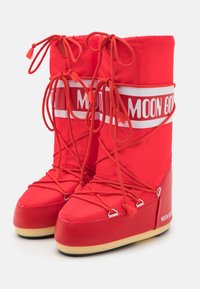 Red high-top Moon Boot snow boots with white logo bands, crisscrossing red laces, and beige soles on a white background.