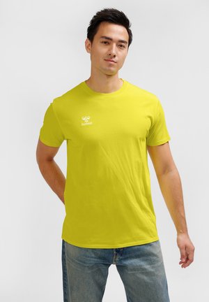 Mężczyzna z krótkimi ciemnymi włosami, ubrany w jaskrawożółtą koszulkę Hummel i niebieskie dżinsy, stojący na jednolitym jasnym tle.