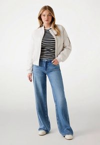 Veste bomber beige clair portée sur un t-shirt rayé noir et blanc, associée à un jean large bleu et des baskets blanches. Texture lisse sur toute la surface.