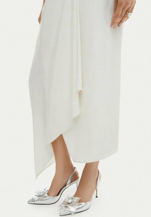 Srebrne szpilki typu slingback z ozdobami w postaci kwiatów i pereł, noszone z lejącą się białą asymetryczną spódnicą.