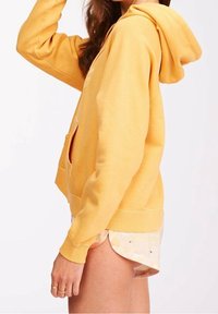 Sudadera amarilla con un bolsillo frontal, cordón ajustable y puños de canalé. Tejido suave, corte casual y costuras en contraste visibles.