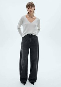 Lichtgrijze V-hals trui gemaakt van zacht materiaal, gecombineerd met hoge, wijde zwarte jeans. Model draagt puntige zwarte hakken.