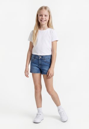 Hymyilevä tyttö, jolla on pitkät vaaleat hiukset ja joka on pukeutunut valkoiseen t-paitaan, sinisiin farkkushortseihin, valkoisiin sukkiin ja valkoisiin lenkkareihin seisten valkoisella taustalla.